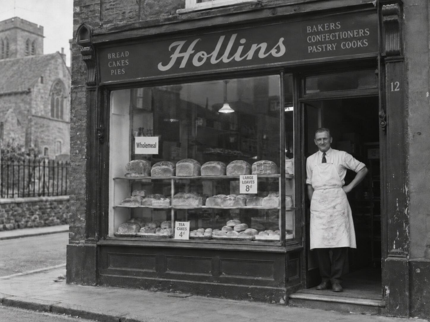 Black-and-white photo of the original Hollins shopfront in 1968, Ted in his apron at the door beside a window full of loaves